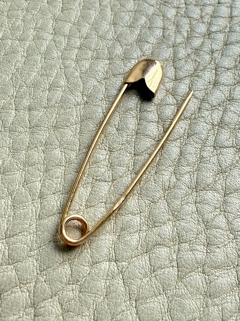 An 18k solid gold safety pin brooch resting on a textured light gray leather surface.