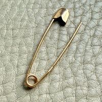 An 18k solid gold safety pin brooch resting on a textured light gray leather surface.