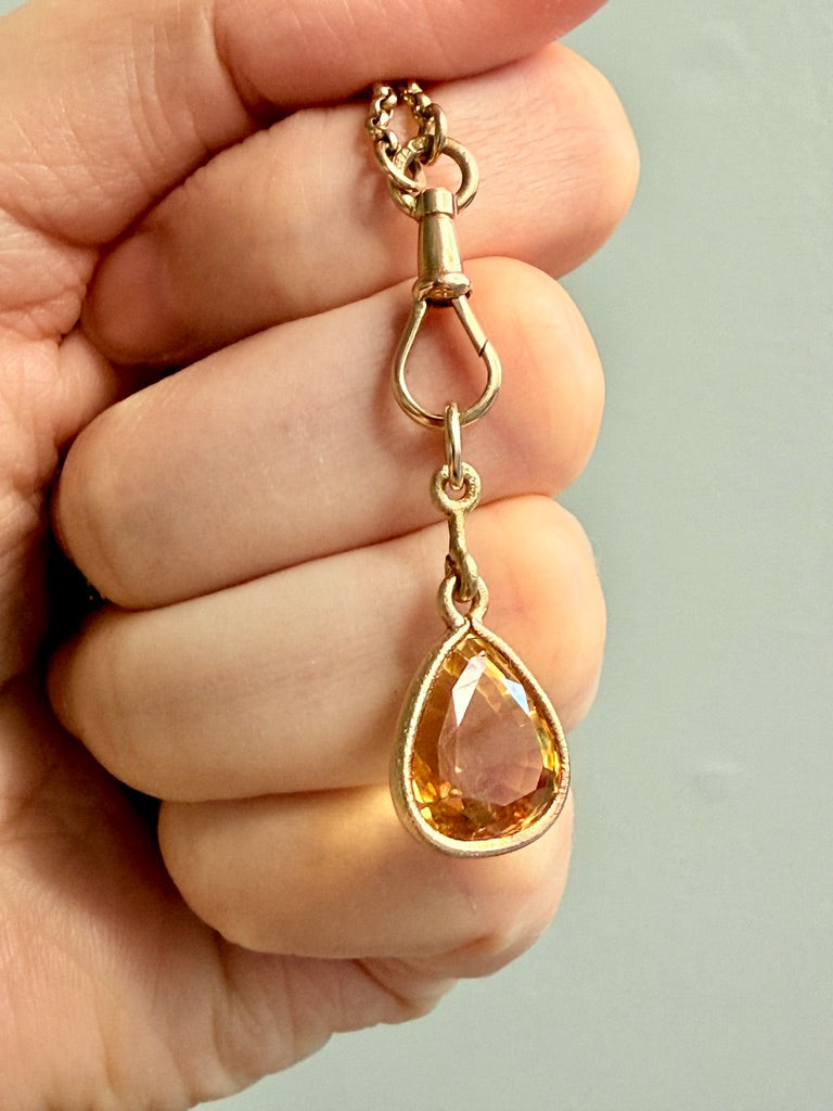 Gold pendant with a teardrop-shaped orange gemstone held in a hand against a neutral background