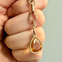 Gold pendant with a teardrop-shaped orange gemstone held in a hand against a neutral background