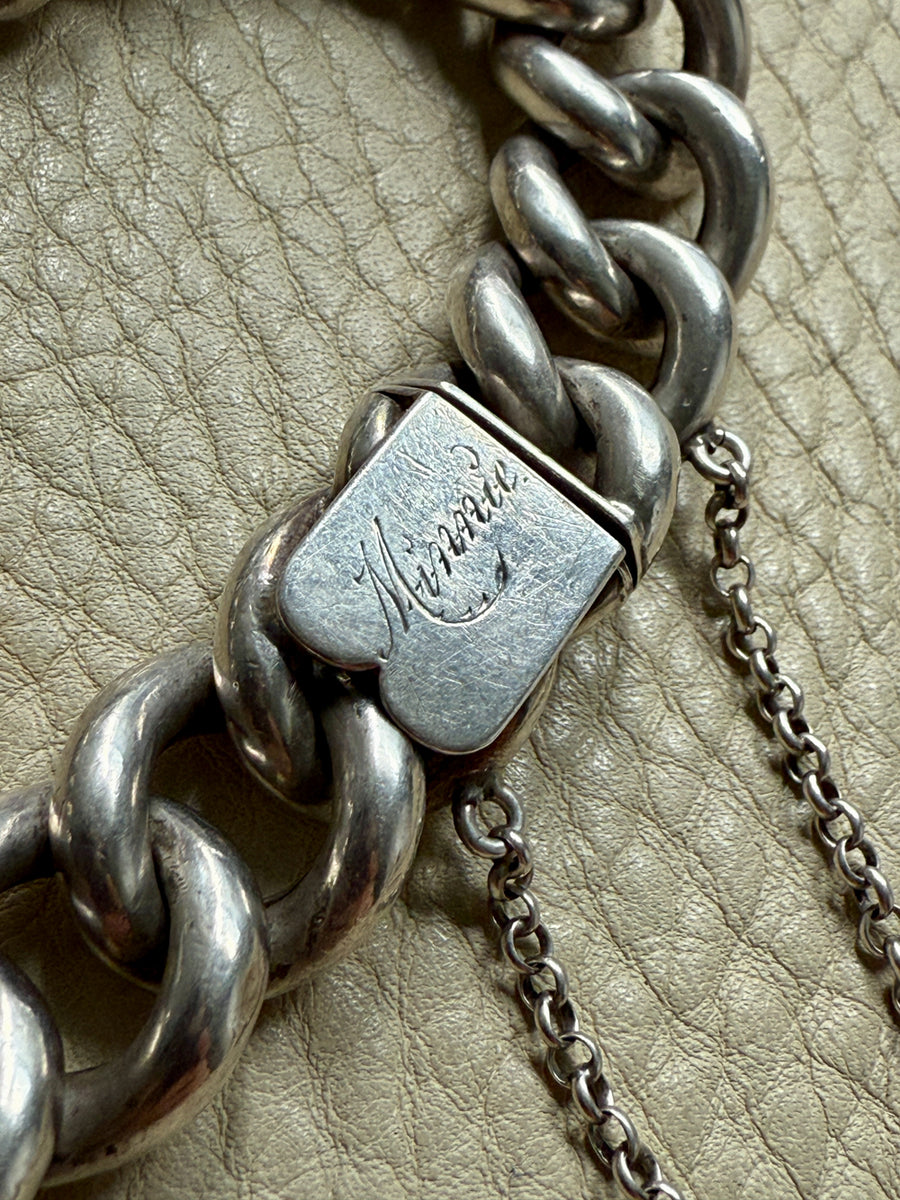 Close-up of a silver chain bracelet clasp with engraving for Minnie, on a beige leather surface