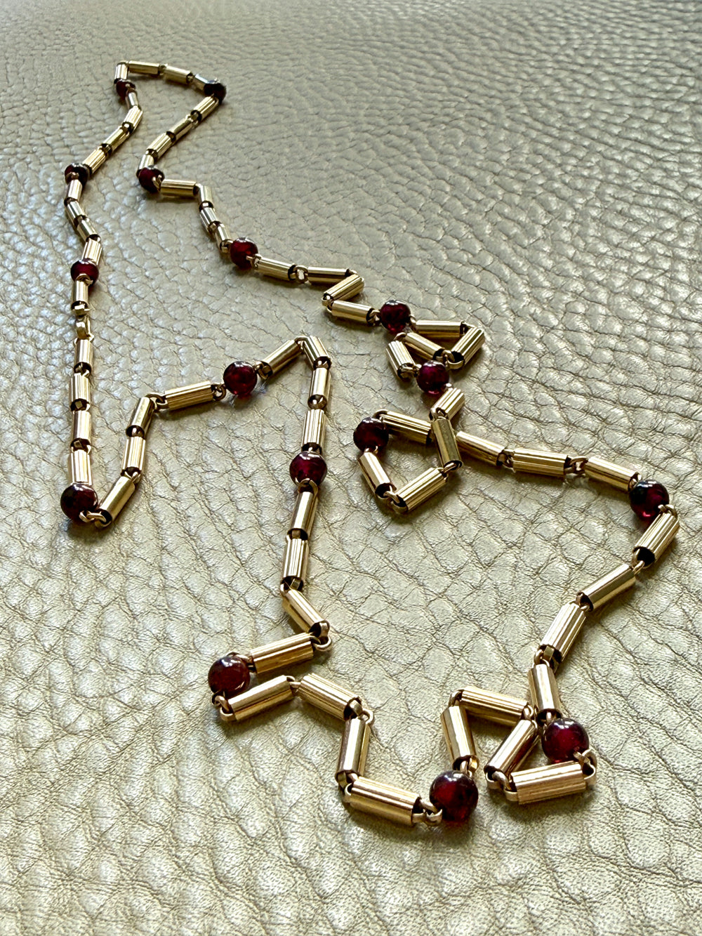 An 18k gold chain necklace with cylindrical links and small dark red gemstone accents, laid out on a textured light beige surface.