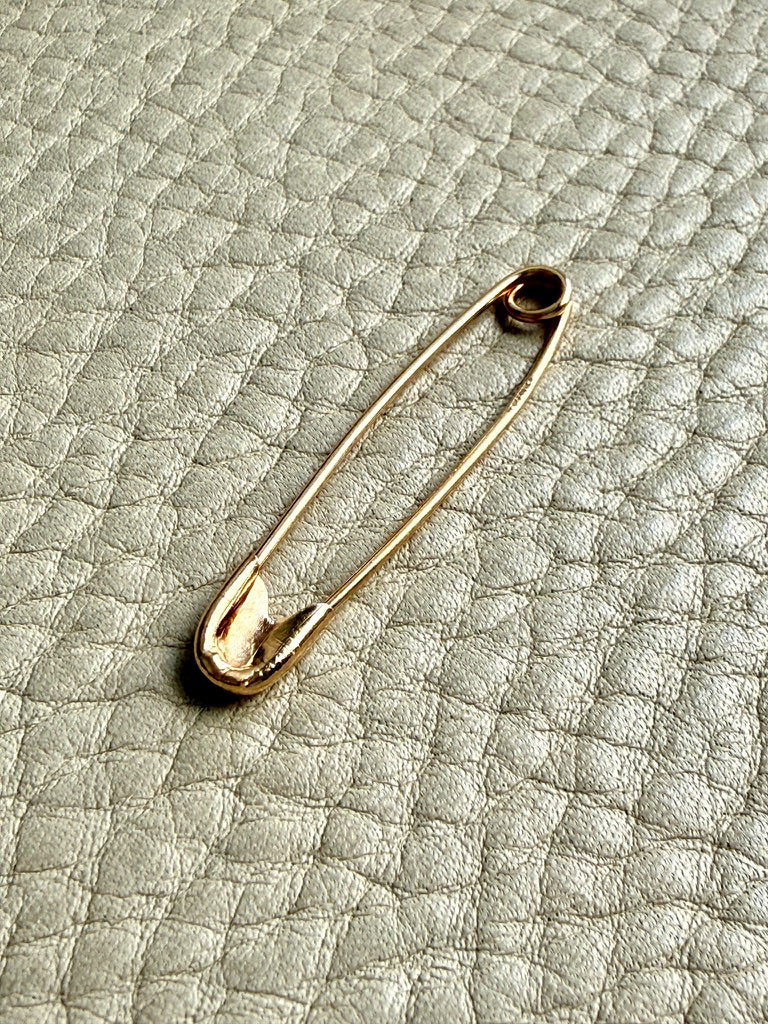 A gold safety pin brooch resting on a textured light gray leather surface.