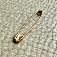 A gold safety pin brooch resting on a textured light gray leather surface.