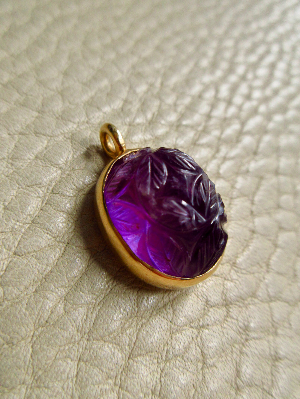 An 18k gold vintage pendant with a carved purple amethyst cabochon, resting on a textured beige leather surface.
