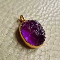 An 18k gold vintage pendant with a carved purple amethyst cabochon, resting on a textured beige leather surface.