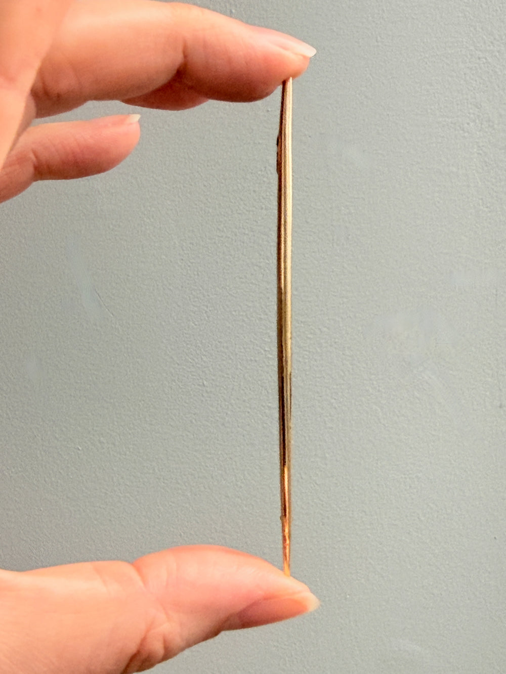 Hand holding a thin, long 18k gold long brooch pin against a plain background