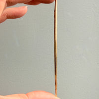 Hand holding a thin, long 18k gold long brooch pin against a plain background
