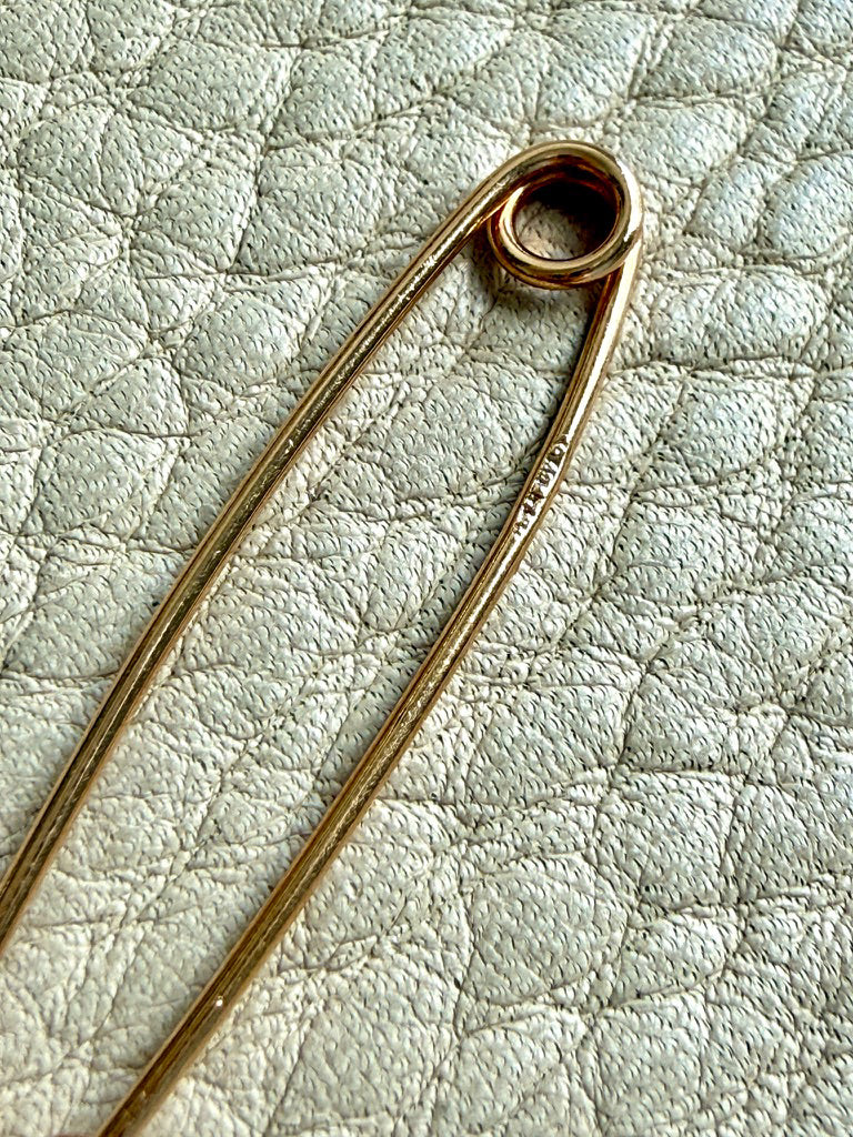 A close-up of a gold safety pin brooch resting on a textured light beige leather surface.