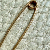 A close-up of a gold safety pin brooch resting on a textured light beige leather surface.
