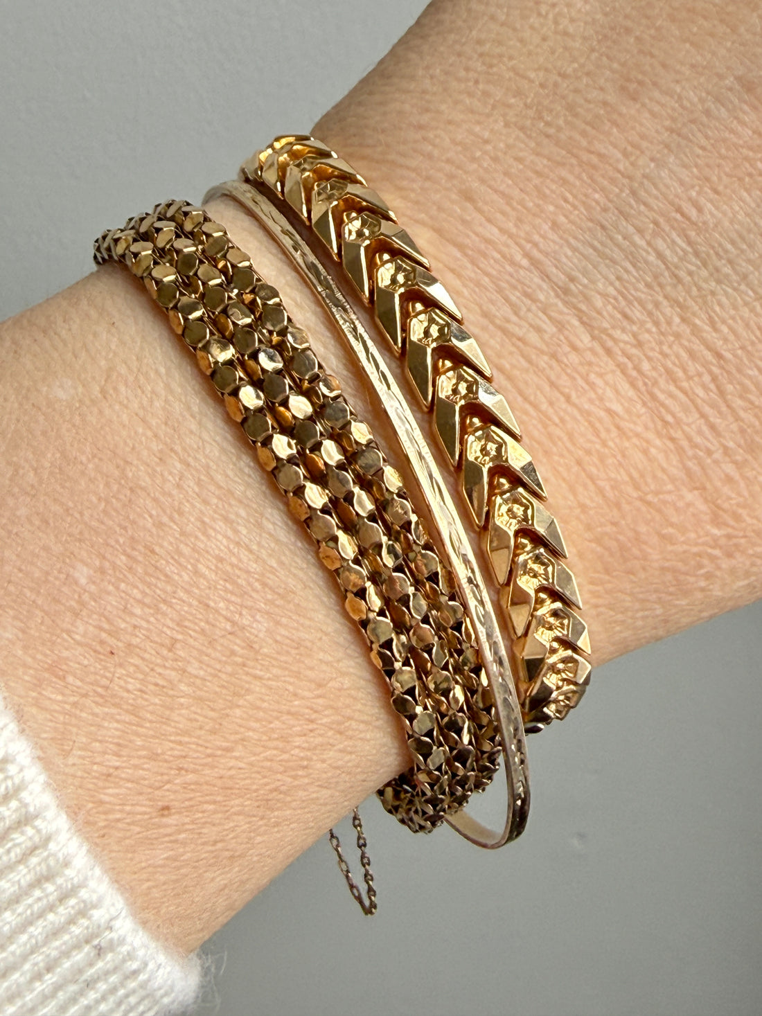 A close-up of a person's wrist wearing a stack of vintage 18k gold bracelets, including a faceted chain bracelet, a textured bangle, a smooth bangle, and a chevron-patterned bangle.