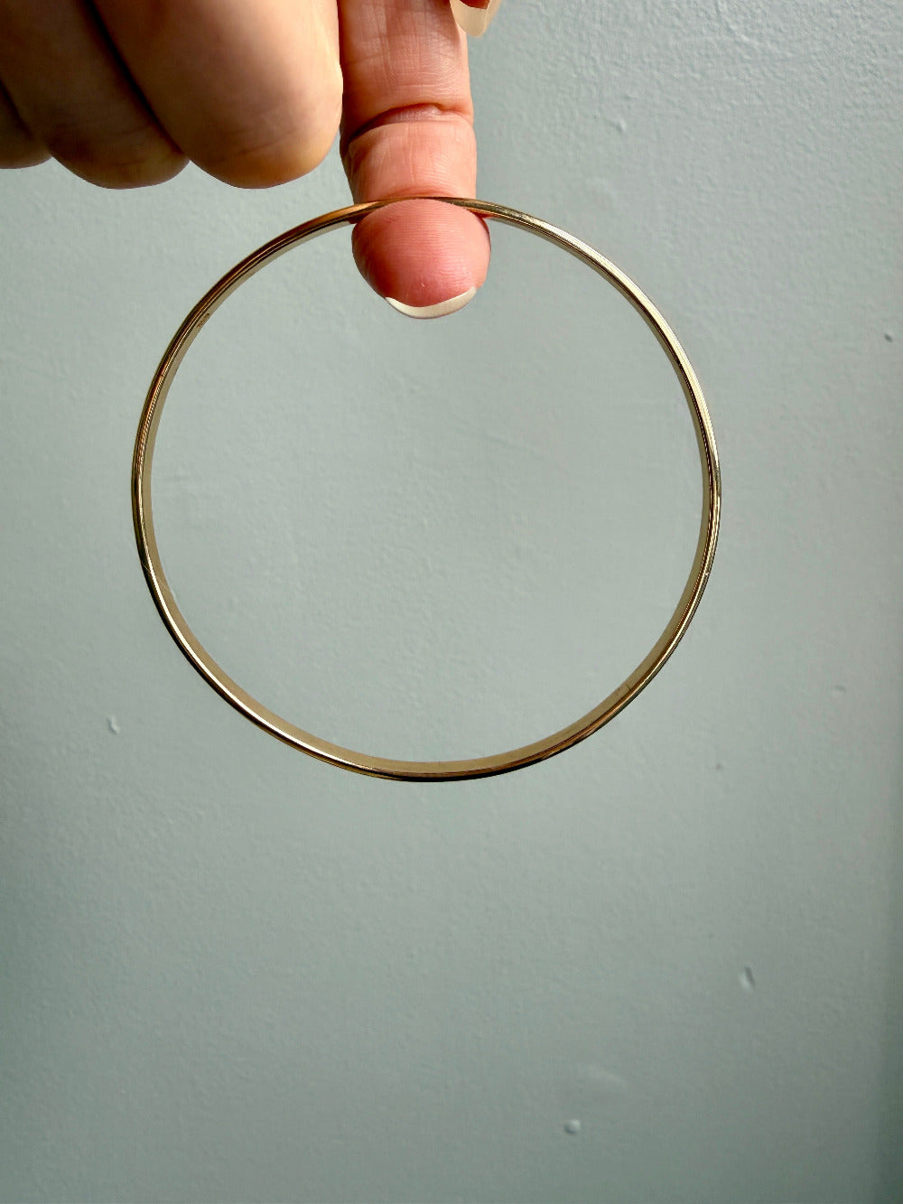 A person's hand holding a narrow, polished gold bangle against a plain, light gray wall.