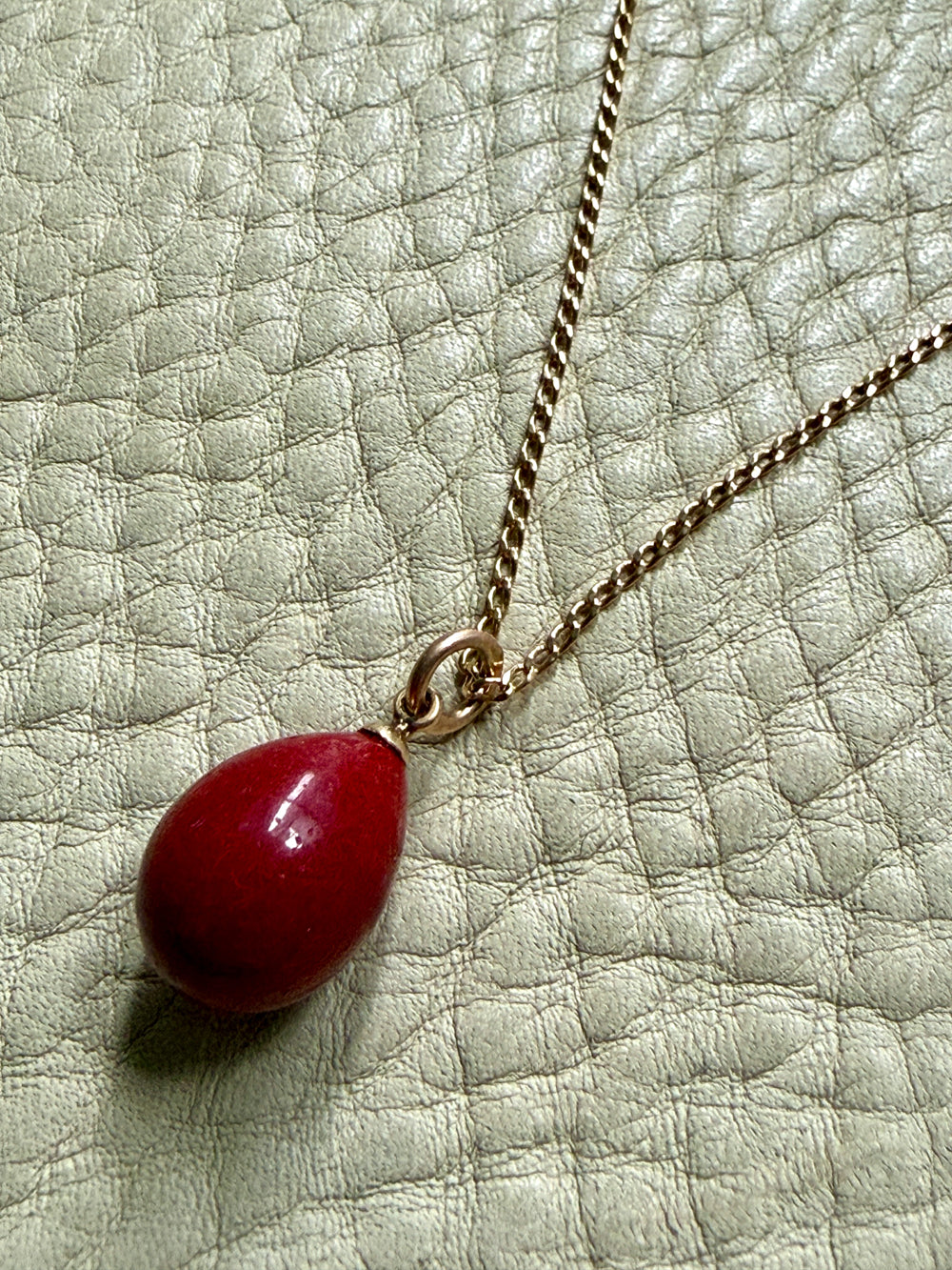red purpurin egg pendant on a gold chain against a textured light green background