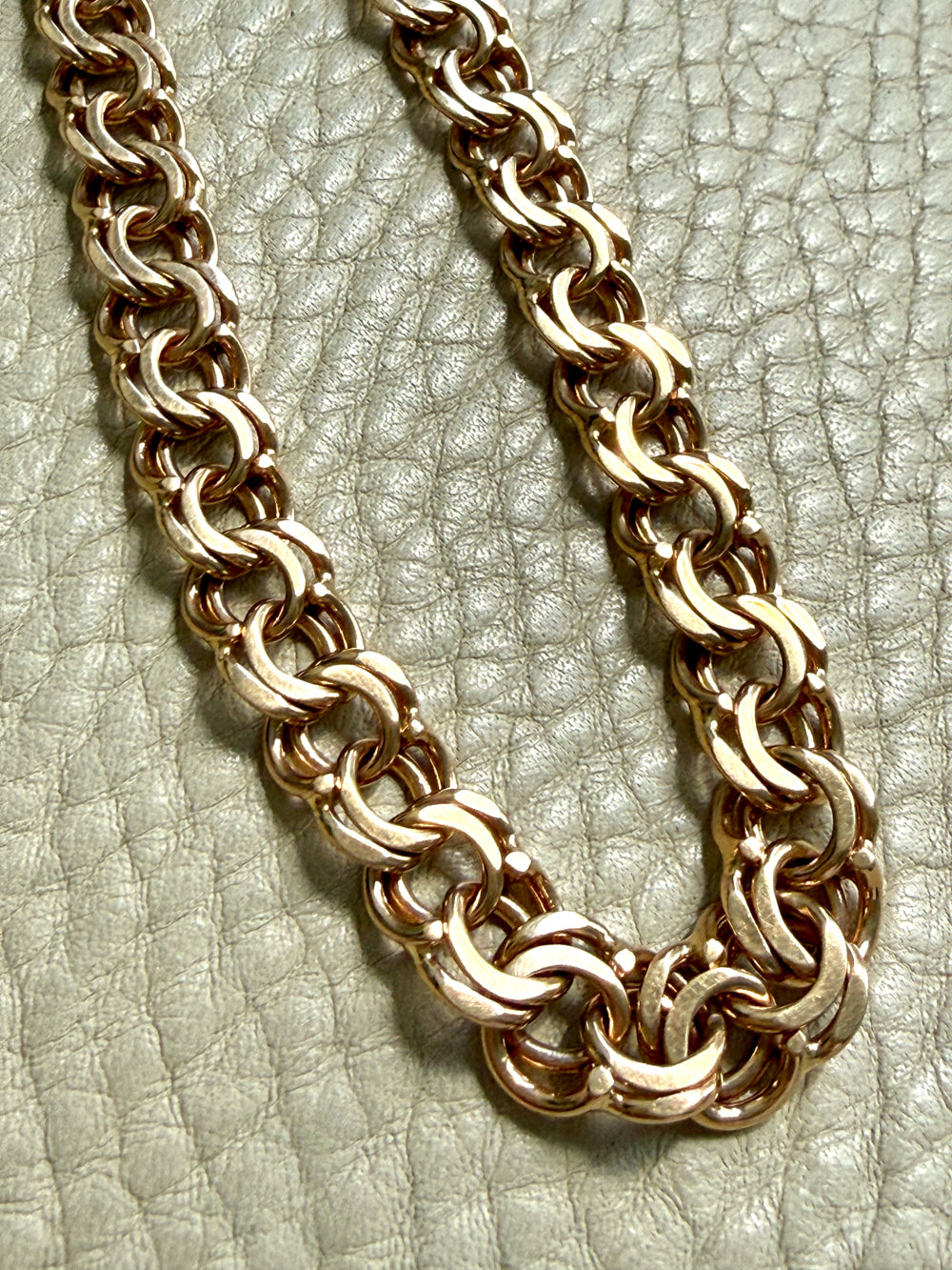 A close-up of a chunky 18k gold chain link necklace resting on a textured light beige leather surface.