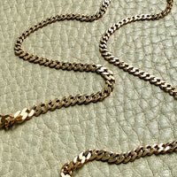 An 18k gold pressed curb link chain necklace draped across a textured light beige leather surface.