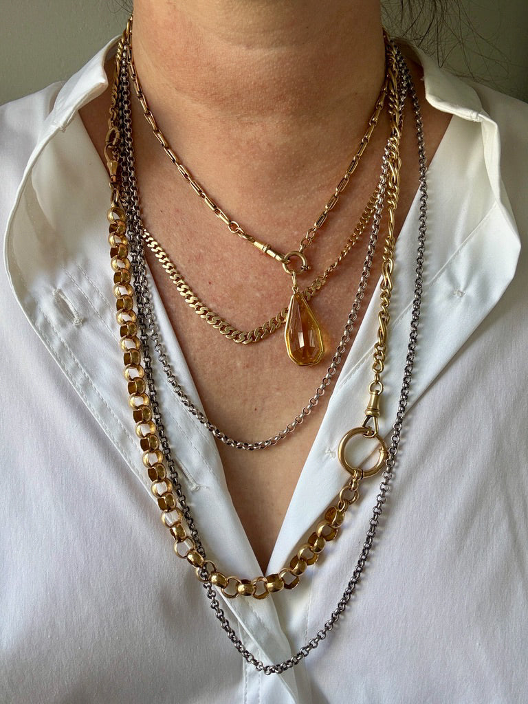 A close-up of a person wearing a layered necklace stack featuring multiple gold and silver chains, including a teardrop pendant, on a white collared shirt.