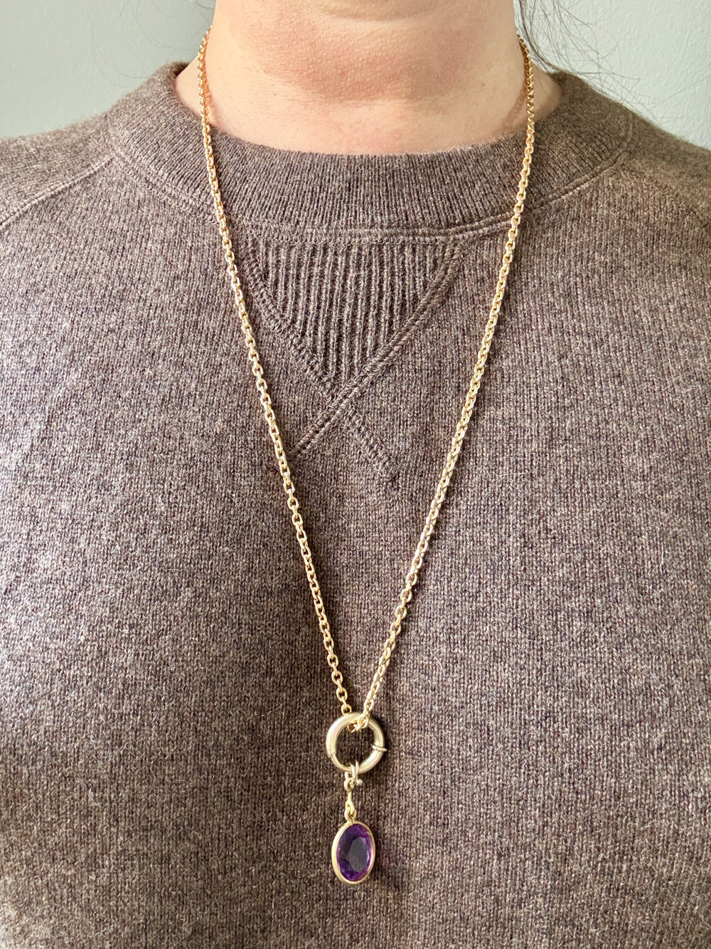 Gold necklace with a purple pendant worn by a person in a brown sweater.