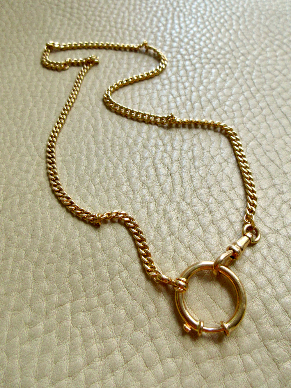 A gold chain necklace with a large circular bolt ring clasp, displayed on a textured beige leather surface. penelopepenelope.com