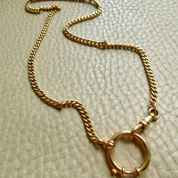 A gold chain necklace with a large circular bolt ring clasp, displayed on a textured beige leather surface. penelopepenelope.com