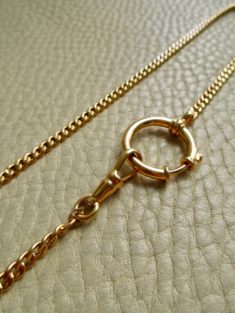 A close-up of an 18k gold chain necklace with a circular bolt ring clasp, laid on a textured beige leather surface.