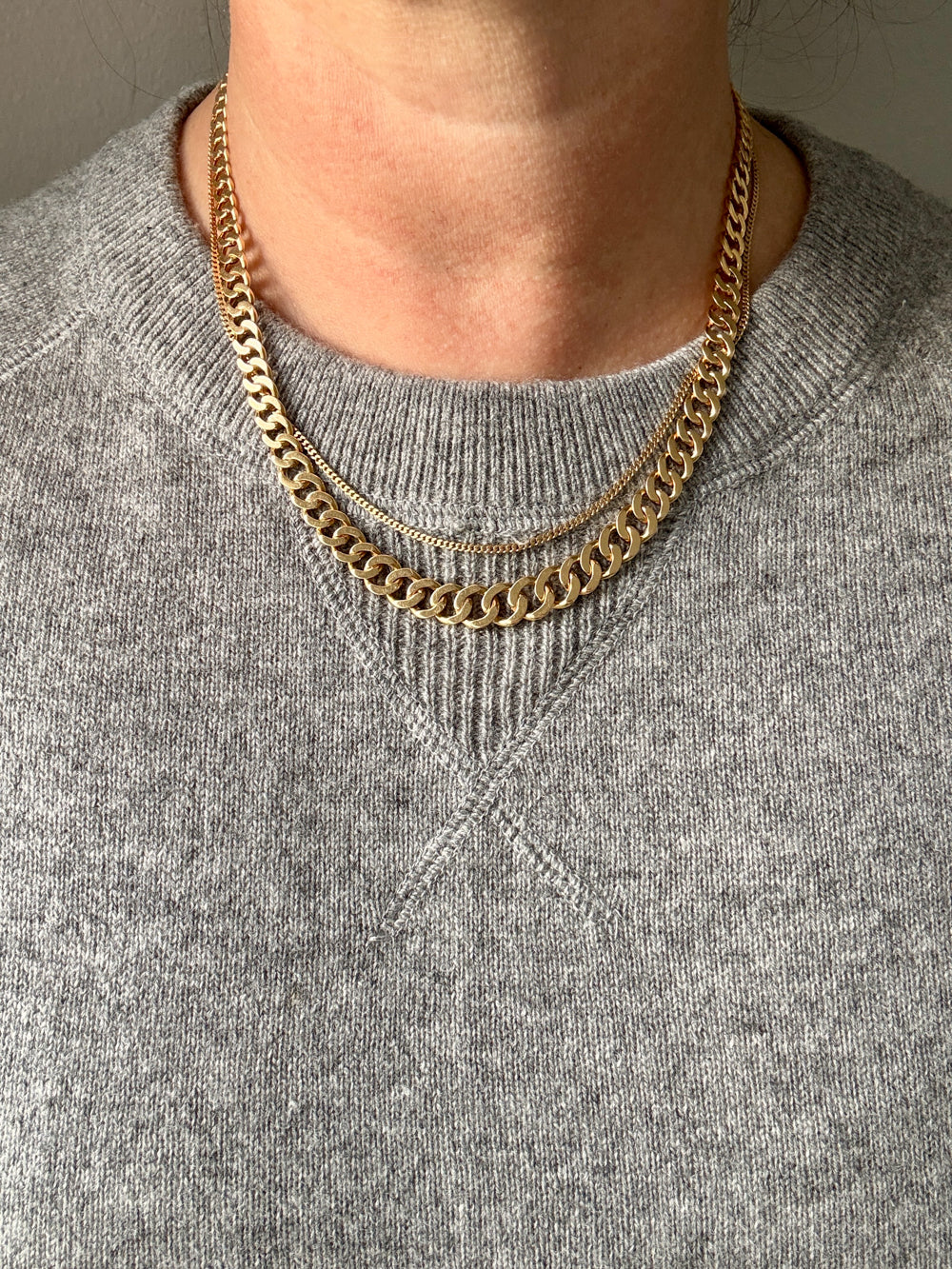 A close-up of a person wearing a double-layered gold chain necklace over a gray knit crewneck sweater.