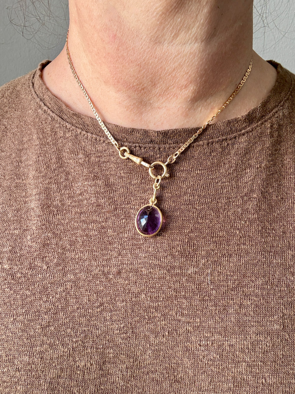 Gold necklace with a purple gemstone pendant worn by a person in a brown shirt.