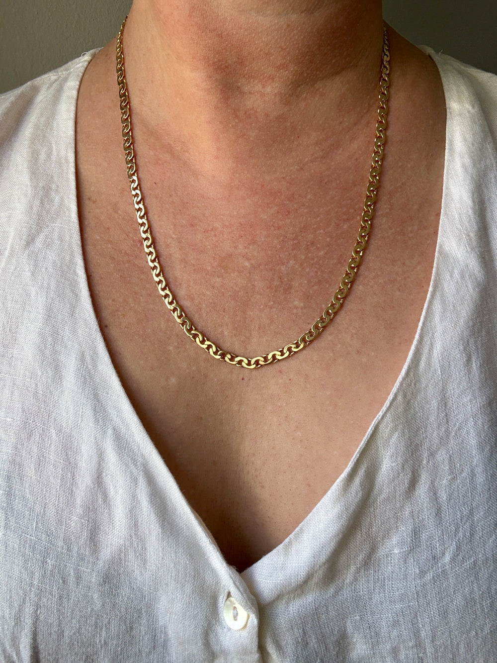 A close-up of a person wearing an 18k gold virola link chain necklace over a white linen V-neck top.