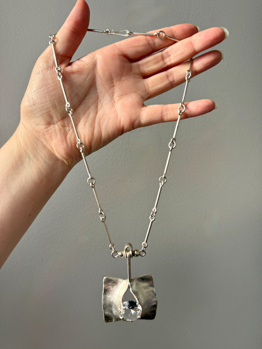 Hand holding a silver necklace with a unique pendant against a neutral background