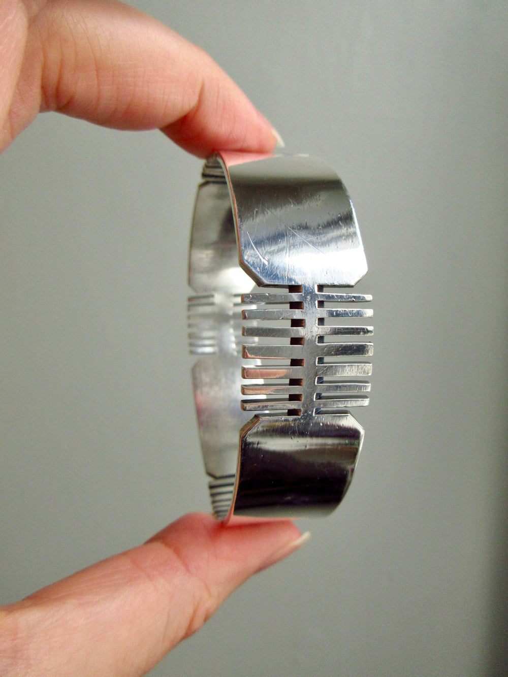 A hand holding a polished silver bangle bracelet with a geometric, ribbed design against a plain gray background.