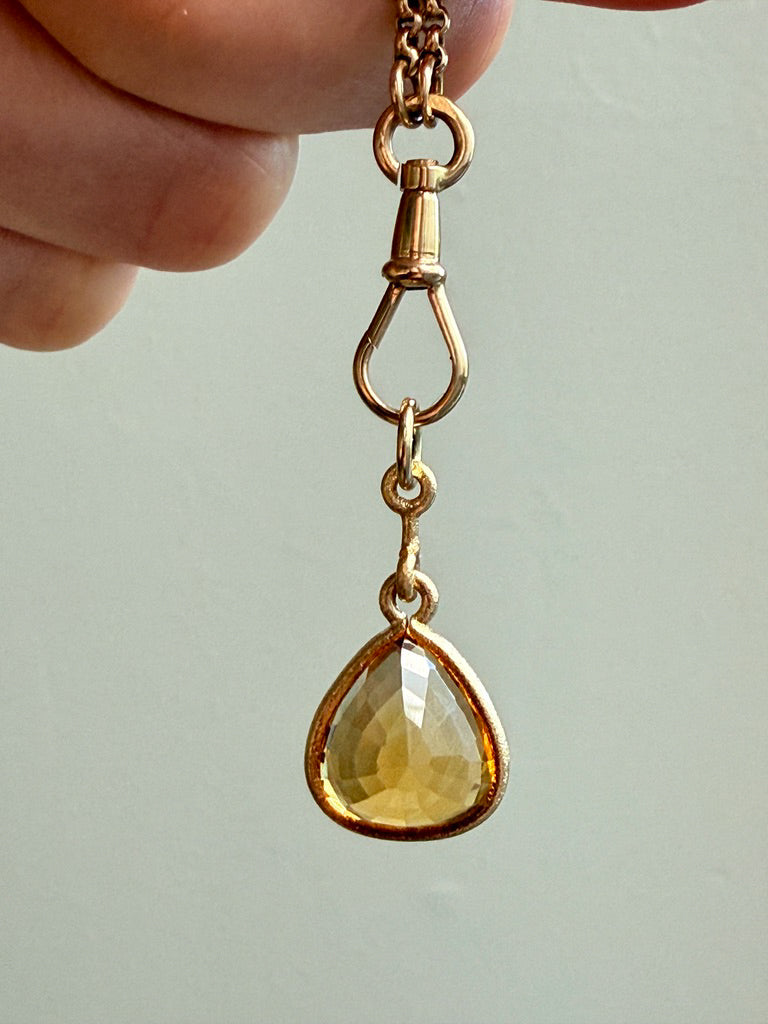 Gold pendant with a teardrop-shaped yellow gemstone held by a hand against a light background