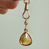 Gold pendant with a teardrop-shaped yellow gemstone held by a hand against a light background