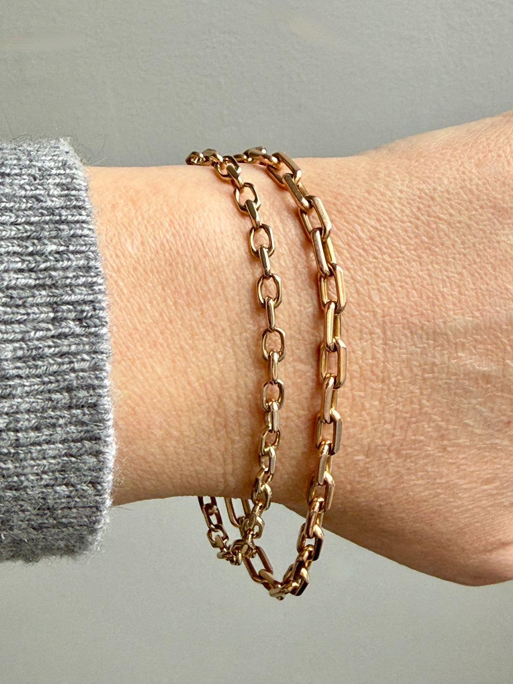 Gold chain bracelets on a wrist with a gray sleeve against a neutral background