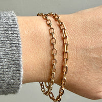 Gold chain bracelets on a wrist with a gray sleeve against a neutral background