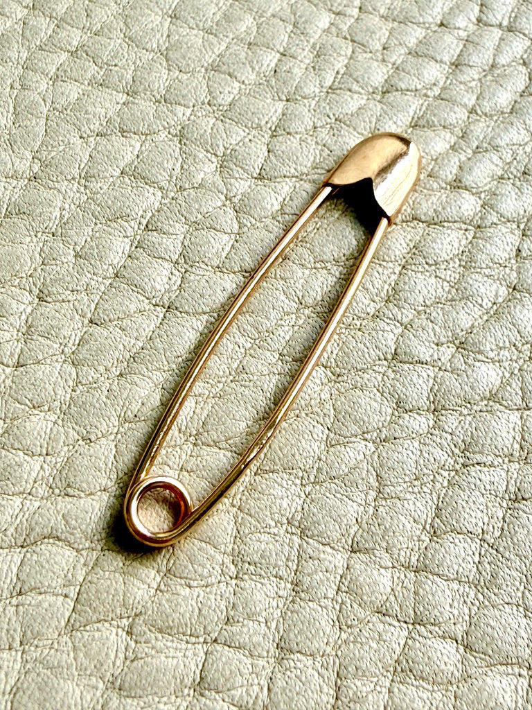 An 18k solid gold safety pin brooch resting on a textured light beige leather surface.