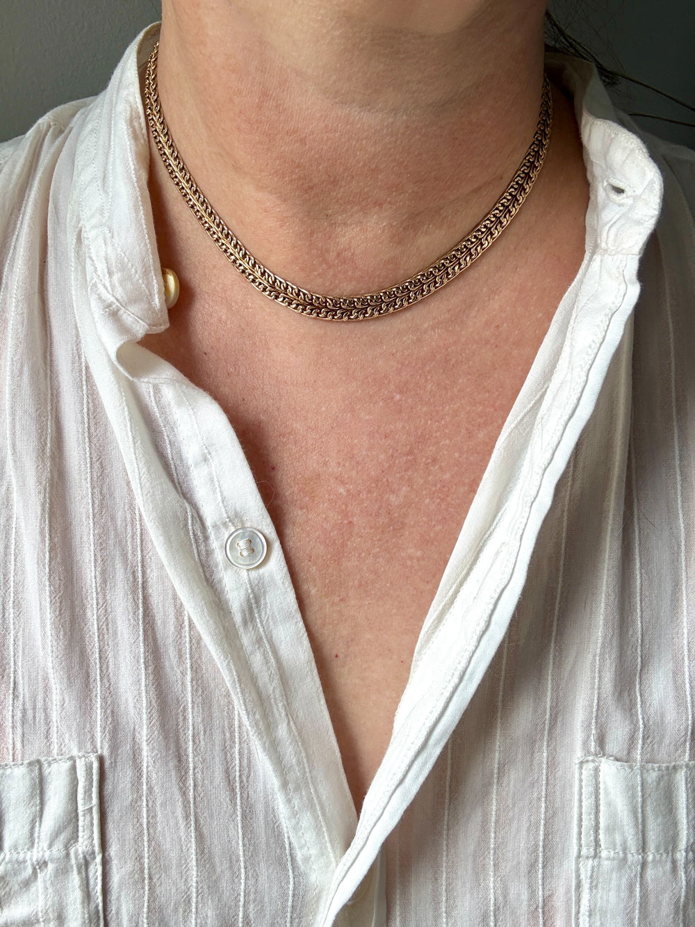 A close-up of a person's neck and upper chest wearing a textured, chain-link style gold necklace over a white button-down shirt.