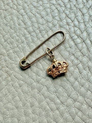 Gold safety pin with a crown charm on a textured light blue background
