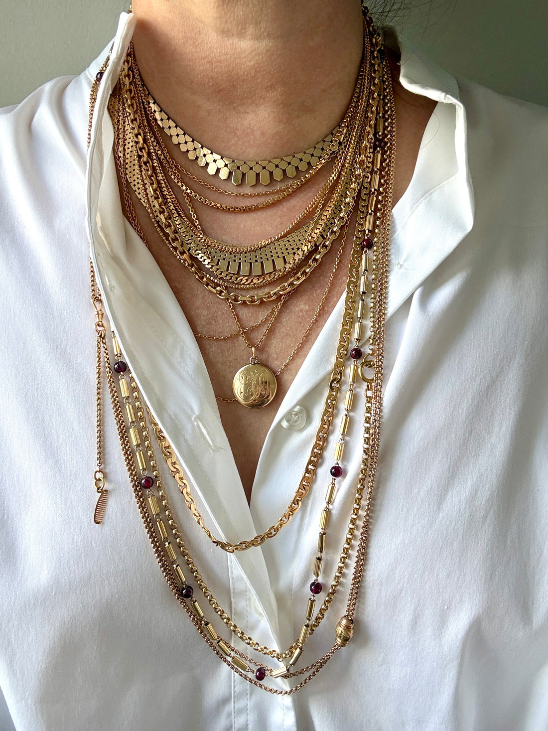 A close-up of a person wearing a layered stack of gold-toned necklaces over a white button-up shirt. The necklaces include chain styles, mesh details, beads, and a round locket pendant.