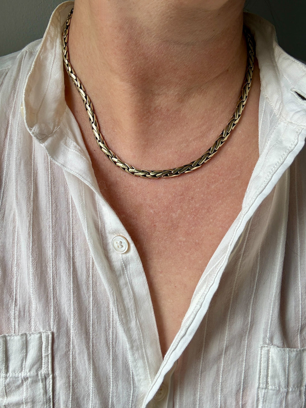 A close-up of a person wearing a braided gold chain necklace over a white button-up shirt.