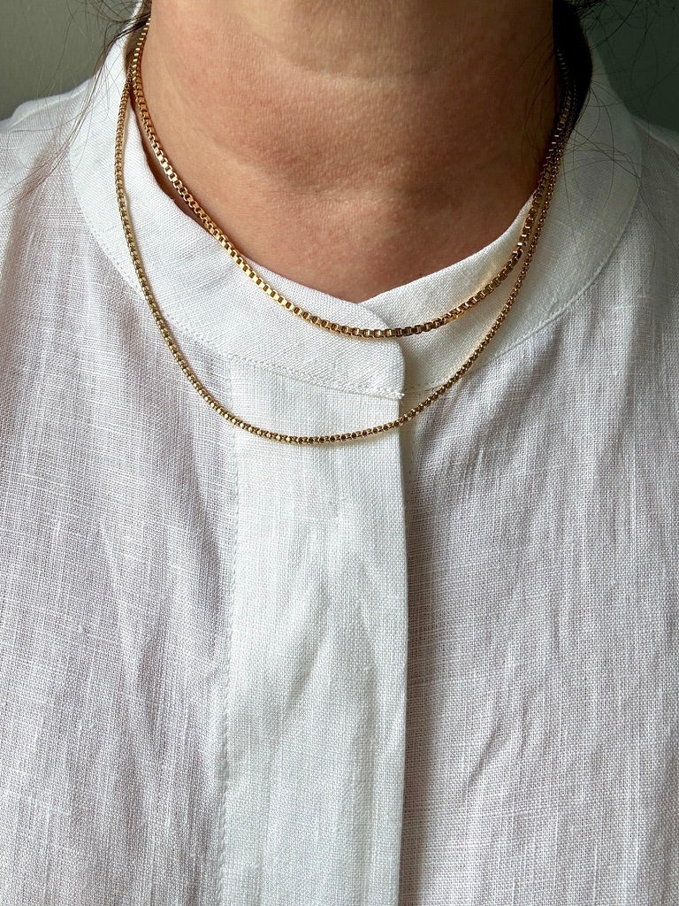 A close-up of a person wearing two gold chain necklaces layered over a white linen shirt.