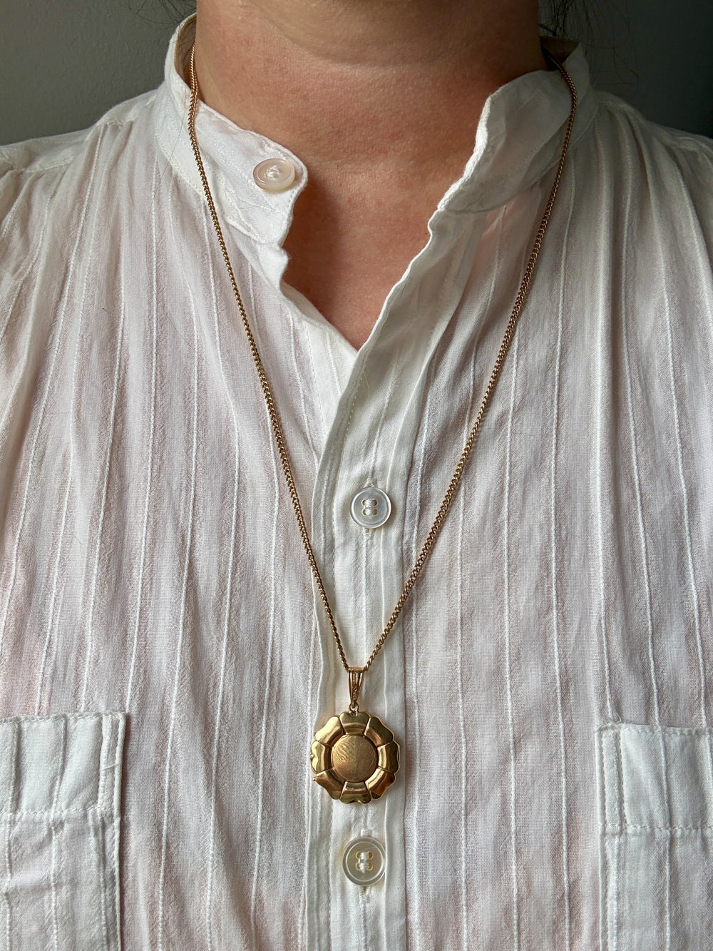 A close-up of a person wearing a gold pendant necklace with a textured circular medallion over a white button-up shirt.