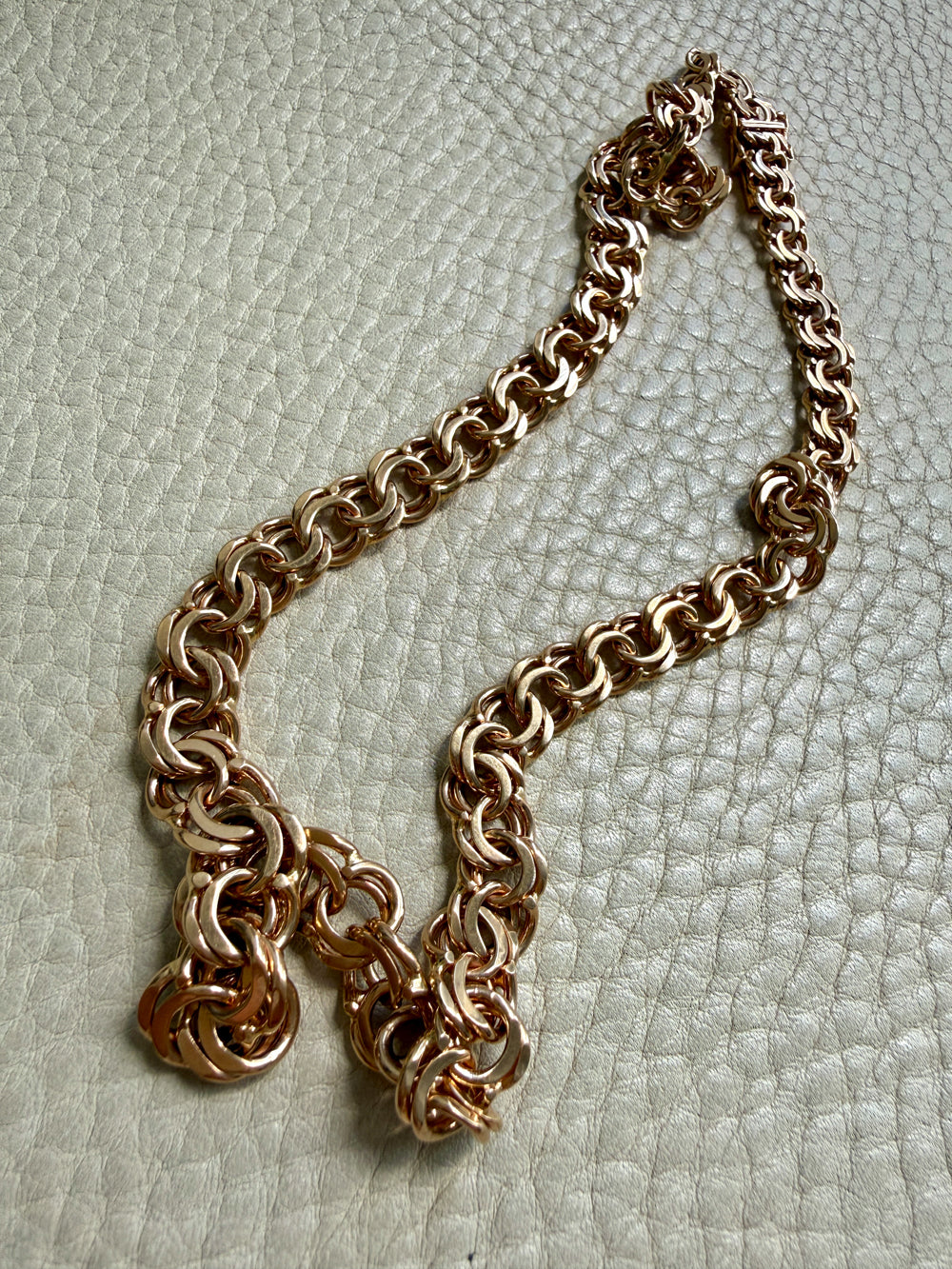 A chunky rose gold chain necklace with a textured, interlocking link design, displayed on a light beige textured leather surface.