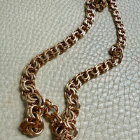 A chunky rose gold chain necklace with a textured, interlocking link design, displayed on a light beige textured leather surface.