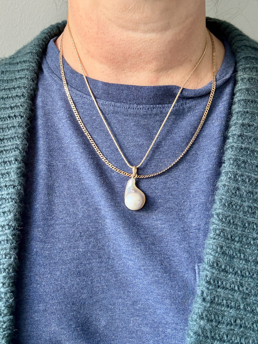 Person wearing a blue shirt and green cardigan with a gold necklace featuring a pearl pendant.