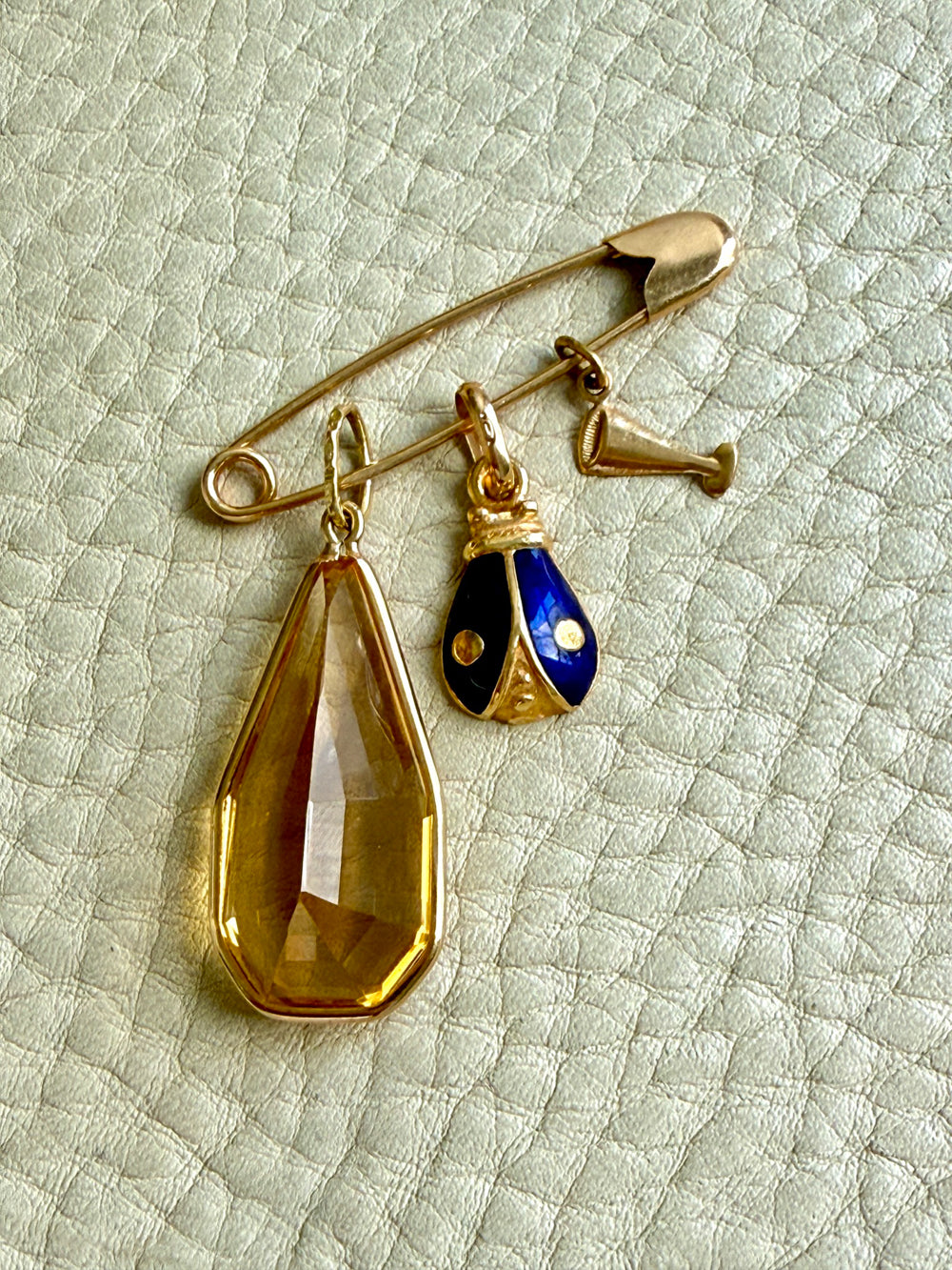 A gold safety pin brooch with three dangling charms: a large faceted amber-yellow teardrop crystal, a small gold and blue enamel ladybug charm, and a small gold cava glass charm, displayed on a textured off-white leather surface.