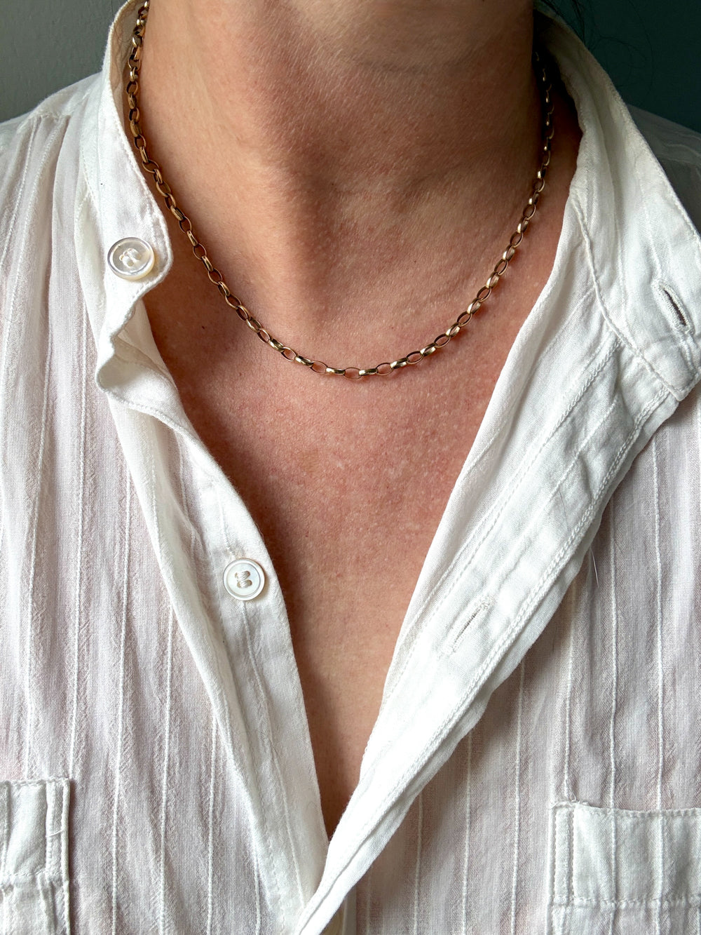 A close-up of a person's neck and chest wearing a delicate 9k gold chain necklace over a white button-down shirt.