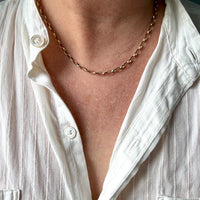 A close-up of a person's neck and chest wearing a delicate 9k gold chain necklace over a white button-down shirt.