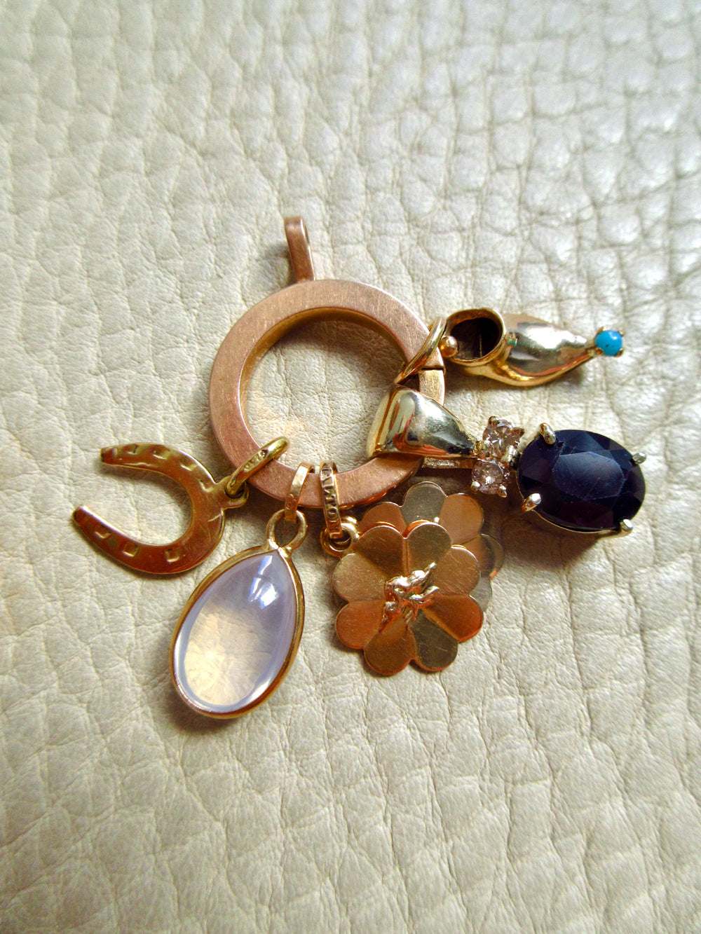 A gold charm cluster featuring a large circular ring, a horseshoe, a flower-shaped charm, a teardrop moonstone pendant, a small bird charm, and a dark oval sapphire gemstone, displayed on a textured beige leather background.