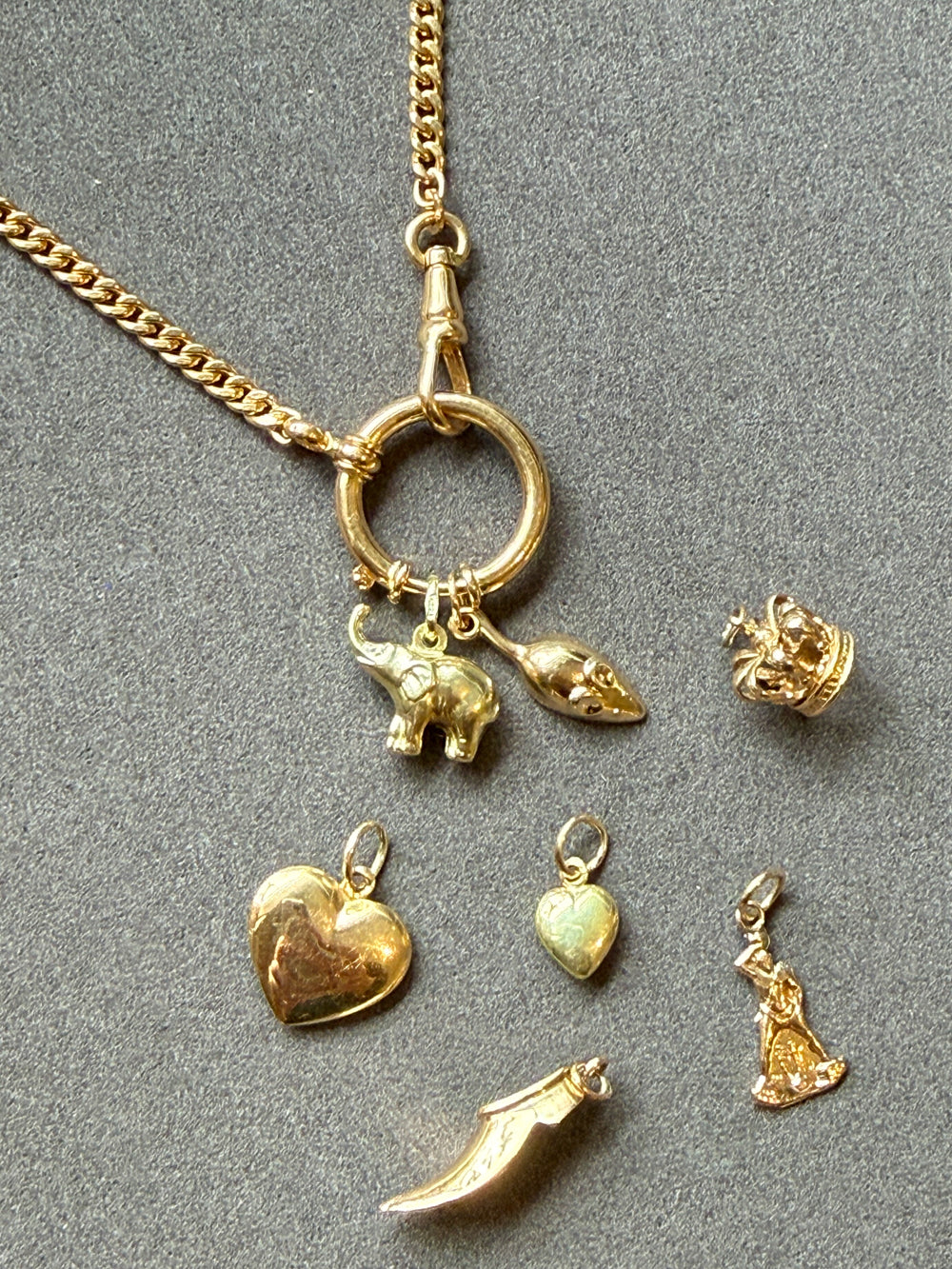 An 18k gold watch chain necklace with a bolt ring clasp and multiple dangling charms, including an elephant, mouse, crown, heart, shoe, and small figurine, arranged on a gray textured background.