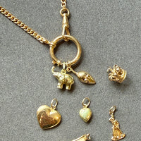 An 18k gold watch chain necklace with a bolt ring clasp and multiple dangling charms, including an elephant, mouse, crown, heart, shoe, and small figurine, arranged on a gray textured background.