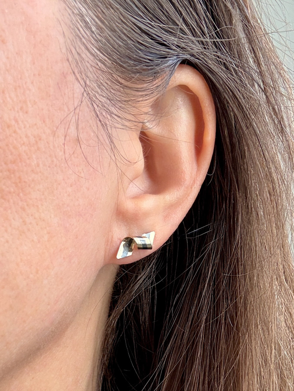 A close-up of a person's ear wearing a small geometric stud Paul Morelli earrings with silver and gold stripes